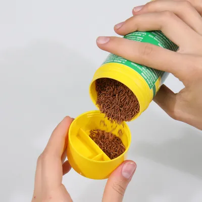 Ruka sype hnědé granule ze žluté dózy se zelenou etiketou do žluté přihrádky s oddíly.