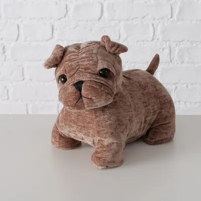 Peluche marrone a forma di cane con occhi neri e coda corta, su un tavolo bianco davanti a una parete di mattoni.