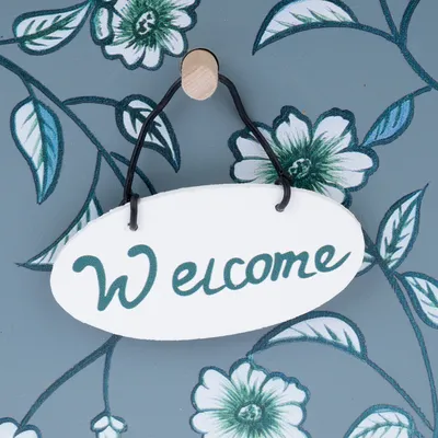 Uvítacia tabuľa z bieleho dreva so zeleným nápisom 'Welcome', zavesená na čiernom drôte pred kvetinovým pozadím.