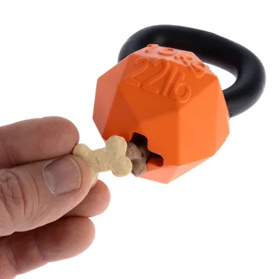 Orangefarvet hundelegetøj i kettlebell-form med sort håndtag, kan fyldes med godbidder. Hånd lægger benformet godbid i åbningen.