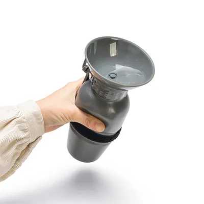 Hand hält graue Trinkflasche mit integriertem Napf, geeignet für unterwegs. Flasche hat einen Klettverschluss zur Befestigung.