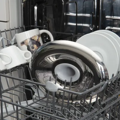 Accessoire en métal et plastique placé dans un lave-vaisselle avec des tasses blanches, dont une avec photo de chien, et des assiettes blanches visibles sur le panier inférieur.