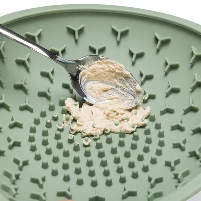 Grüne Silikonmatte mit erhöhten Noppen, darauf wird mit einem Löffel eine cremige Paste verteilt.