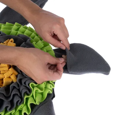Hände zeigen, wie ein Leckerli in eine Tasche aus grauem Stoff gesteckt wird. Umgeben von grünem und gelbem Stoff mit Rüschenstruktur.