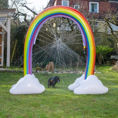 Oppustelig regnbue-vandsprinkler med hvide skyer, vandstråler i alle retninger. Hund i haven løber gennem sprinkleren.