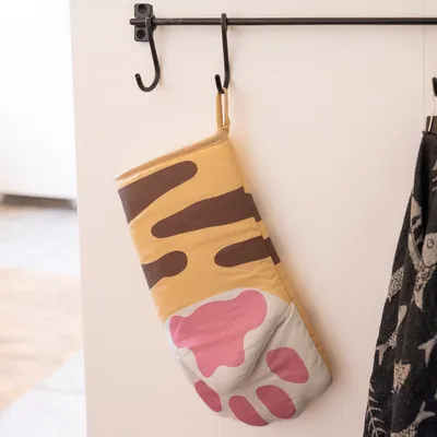 Chňapka se žlutým a hnědým vzorem, růžový design otisku tlapky, zavěšená na černém háčku.