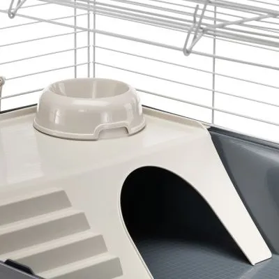 Interno di una gabbia per piccoli animali con ciotola in plastica, scaletta e rifugio integrato. Struttura in metallo e plastica visibile.