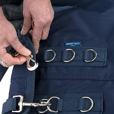 Nomad Tales hondentuig in donkerblauw met meerdere metalen ringen en clipgesp. Handen tonen het vastmaken van de clip aan het tuig.