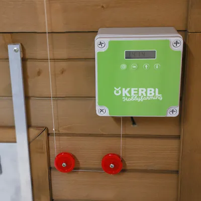 Kerbl Hobbyfarming unitate de control cu afișaj verde, afișare oră și butoane de operare. Două role de deviere roșii vizibile pe suprafață de lemn.