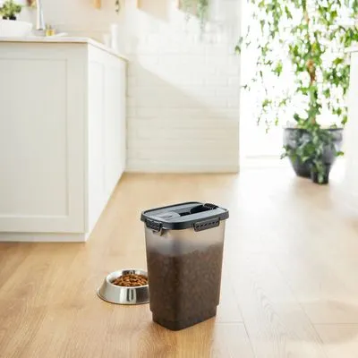 Transparante kunststof voercontainer met zwart deksel, gevuld met brokken. Naast de container staat een metalen voerbak met brokken op een houten vloer in een keuken.