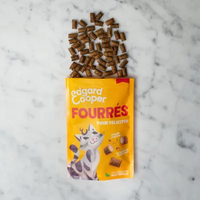 Edgard Cooper Fourrés Pour Féliciter. Gele verpakking met kattenmotief, gevuld met natuurlijke snacks van vers vlees. Kleine bruine traktaties zichtbaar op marmeren tafel.