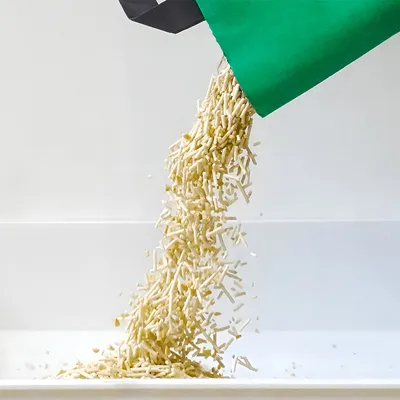Pellet litter being poured from a green bag into a tray, showing product texture and usage method.