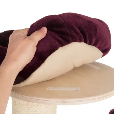 Mână ridicând o pernă textilă bordeaux de pe o platformă rotundă din lemn, cu benzi velcro vizibile pentru fixare. Material: lemn și material textil.