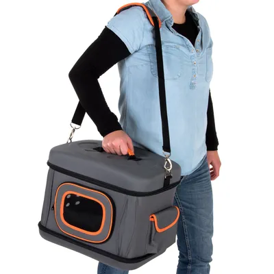Personne portant une caisse de transport grise à bordures orange avec poignée et bandoulière, poche latérale visible, fenêtre grillagée sur le côté.