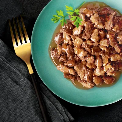 Närbild av saftiga köttbitar i sås, serverade på en turkos tallrik med persiljegarnityr och en gyllene gaffel bredvid.