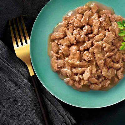 Närbild av saftiga köttbitar i sås på en grön tallrik, dekorerad med persilja. Guldgaffel och svart servett bredvid.