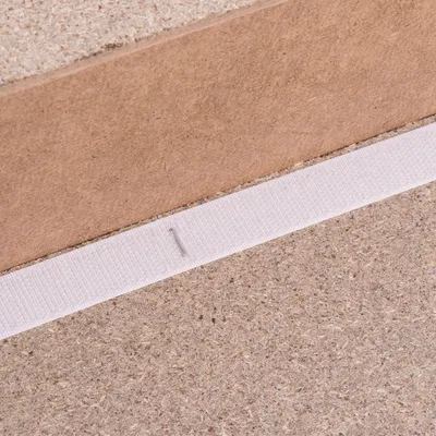 Detail van wit textielband bevestigd met nietje op spaanplaat, zichtbaar tegen achtergrond van bruin houtmateriaal. Materiaal: textiel en spaanplaat.