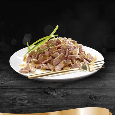 Tallerken med stykker af vådfoder i gelé, pyntet med grønne strimler. Guldgaffel og -kniv ved siden af på sort træbord. Ingen synlig emballage eller mærke.