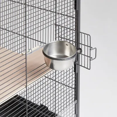 Metall-Futternapf an Gitterkäfig befestigt, mit Holzplattform im Inneren des Käfigs sichtbar.