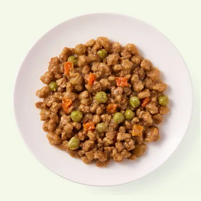 Nahaufnahme eines Tellers mit Nassfutter, bestehend aus braunen Fleischstückchen, grünen Erbsen und orangefarbenen Karottenwürfeln.