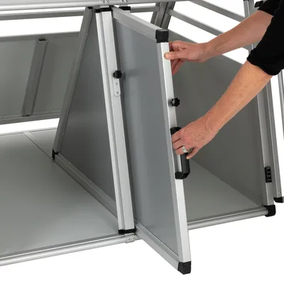 Persona che apre la porta di un trasportino per animali in metallo e pannelli grigi, con maniglia nera visibile.