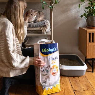 Biokat's Classic 3in1 kattenbakvulling, vrouw naast kattenbak en kat op plank. Verpakking toont hygiënebelofte en productdetails.
