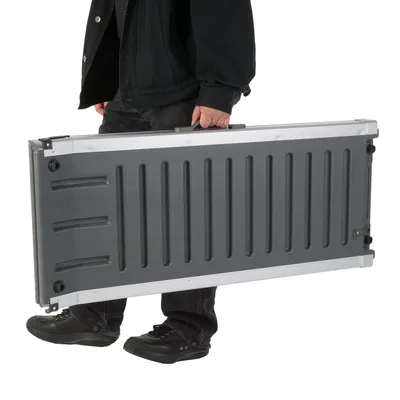 Opgevouwen grijze draagbare kunststof en metalen hondenloopplank met handvat, gedragen door persoon in zwarte kleding en schoenen. Product is rechthoekig en compact.