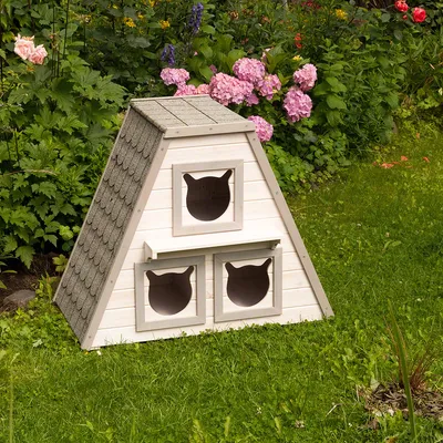Driehoekig wit houten huis met drie kattenvormige ingangen, geschikt voor de tuin. Dak met grijze shingles, omgeven door bloemen en gras.