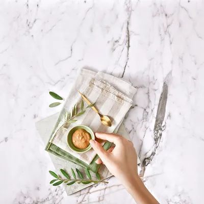 Hand hält eine kleine grüne Schale mit cremigem Inhalt, daneben goldener Löffel und Olivenzweig auf gestreiftem Stofftuch.