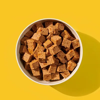 Bowl filled with brown, cube-shaped pet food or treats displayed on a yellow background.