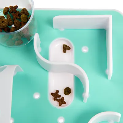 Brinquedo interativo para comida com design de labirinto, base verde e obstáculos brancos. Pedacinhos de ração seca de várias formas visíveis.