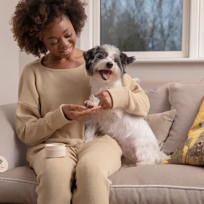 Persoon houdt de poot van een kleine hond vast, naast een blik met verzorgingsproduct op een bank.