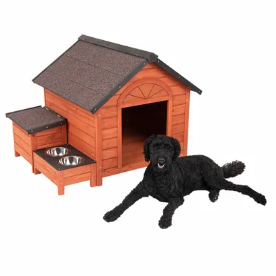 Niche en bois avec toit incliné, deux gamelles en métal intégrées sur le côté et un chien noir allongé devant l'entrée ouverte de la niche.