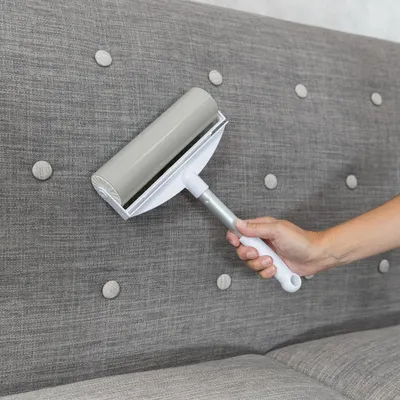 Rouleau adhésif gris et blanc avec manche, utilisé sur un canapé en tissu gris pour enlever les poils ou saletés. Manche en plastique, rouleau large visible.