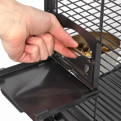 Main ouvrant une trappe métallique sur une cage, accès à une gamelle en métal contenant de la nourriture pour oiseaux ou petits animaux.