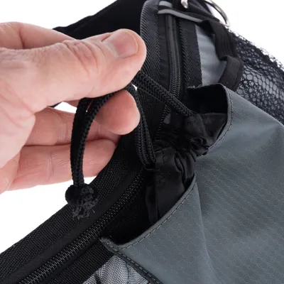 Gros plan sur une main tenant un cordon de serrage noir d’un sac en tissu gris avec poche et filet visible, fermeture éclair noire sur le dessus.