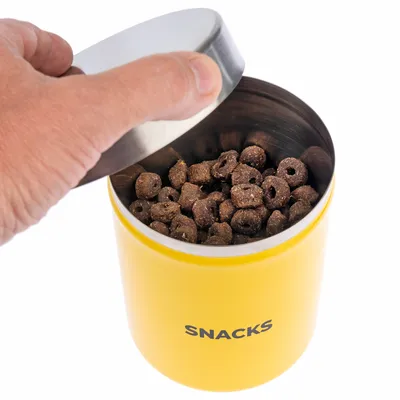 Gelbe Dose mit der Aufschrift 'Snacks', geöffnet, gefüllt mit braunen Leckerlis. Hand hebt den silbernen Deckel an.
