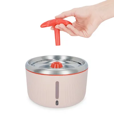 Fontaine à eau pour animaux TRIXIE, bol rond avec couvercle en métal et accessoire rouge en forme de champignon tenu par une main, indicateur de niveau d’eau visible.