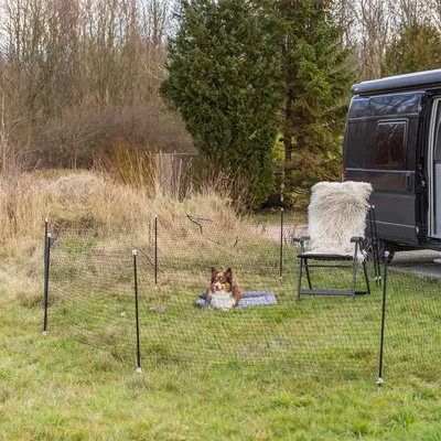 Clôture de jardin en filet noir avec piquets, installée sur l’herbe autour d’un chien couché sur un tapis, à côté d’un camping-car et d’une chaise pliante.