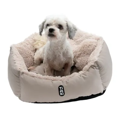 Petit chien blanc assis dans un panier moelleux beige avec intérieur en tissu peluche, marque visible : TIAKI sur l’avant du panier.