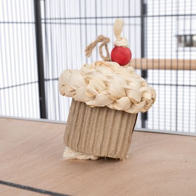 Jouet en forme de cupcake pour oiseaux, composé de carton ondulé, corde tressée et boule en bois rouge, posé dans une cage à barreaux métalliques.