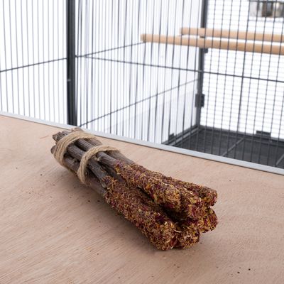 Lot de cinq bâtonnets en bois naturel, extrémités recouvertes d’un mélange végétal séché, attachés avec une ficelle, posés devant une cage pour oiseaux.