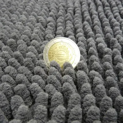 Gros plan sur une pièce de monnaie posée sur un tapis à bouclettes épaisses en tissu gris, montrant la texture et l’épaisseur du matériau par rapport à la taille de la pièce.