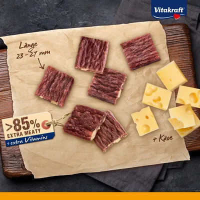 Vitakraft, snacks carrés à la viande longueur 23–27 mm, morceaux de fromage visibles, texte : '>85% extra meaty + extra vitamins', '+ Käse' (fromage en allemand).
