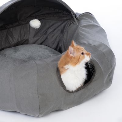 Niche grise en tissu avec coussin intérieur, ouverture ronde sur le côté et balle suspendue, chat roux et blanc visible à l'entrée.