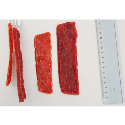 Lamelles de friandises rouges pour animaux posées à côté d'une règle graduée en centimètres, montrant la longueur des morceaux. Fourchette visible à gauche.
