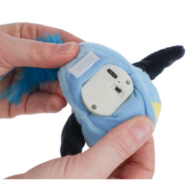 Pluszowa zabawka z wbudowanym modułem elektronicznym, pokazana z przełącznikiem i portem ładowania. Ręce trzymają zabawkę, demonstrując funkcje.