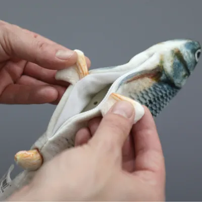 Hand öffnet Reissverschluss eines Fisch-förmigen Spielzeugs, zeigt Innenfach für Zubehör oder Füllung.