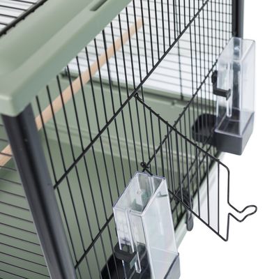 Cage à oiseaux avec grille en métal noir, base en plastique vert, deux distributeurs de nourriture transparents et une perche en bois. Porte ouverte visible.