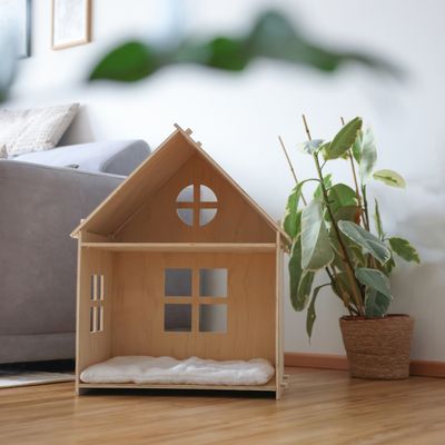 Niche pour animal en bois clair avec coussin blanc, forme de maison avec fenêtres découpées. Placée à l’intérieur près d’un canapé et d’une plante en pot.